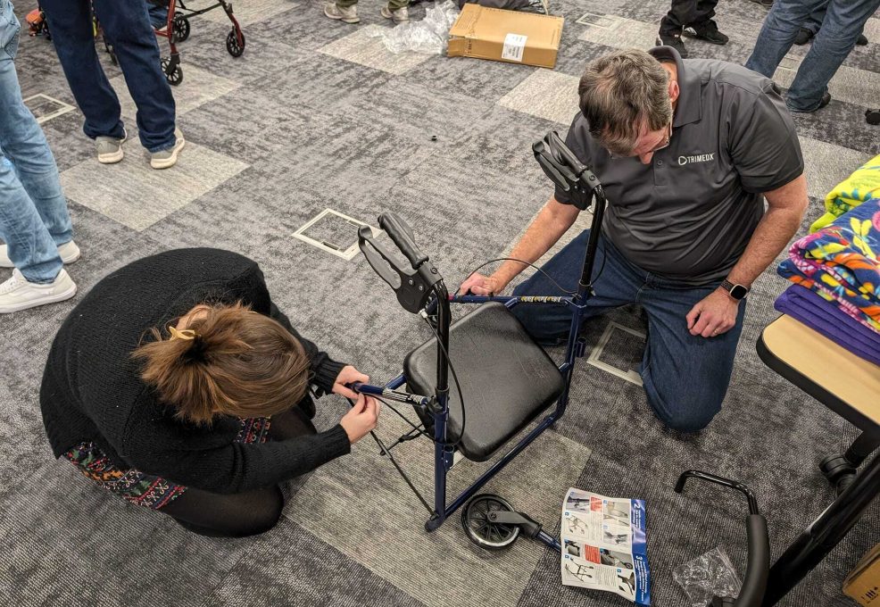 Two participants are kneeling on the floor, carefully assembling a rollator walker by attaching its handles and frame components during a charitable team building activity.