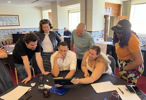 Team building activity focused on human skills training. A group gathers around a tablet, collaborating and strategizing to complete a challenge, reinforcing teamwork, communication, and problem-solving skills.