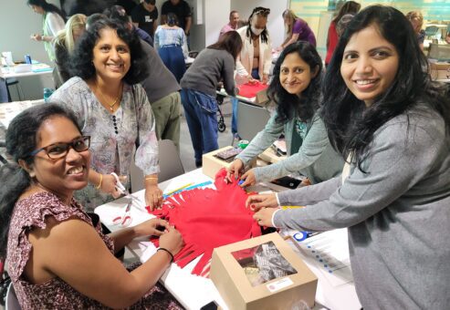 A group of four women smiling while working together on a no-sew fleece blanket, tying knots during a charitable team building event.