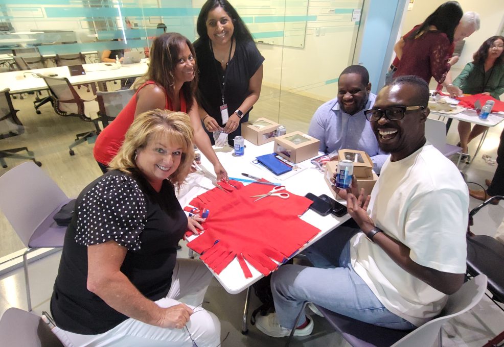 Five smiling participants sit around a table in a brightly lit room, engaged in assembling items for the Rollator Walker Build team building program. On the table are red fabric pieces, scissors, and small boxes, as they work on decorations and assembling parts. The group looks cheerful as they work together.