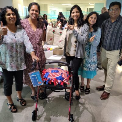 A group of five individuals, smiling and showing a thumbs up, stands around a rollator walker they have built. The walker features an American flag-themed cushion attached to it. One of the women is holding up a canvas tote bag decorated with colorful designs. Various supplies and items are visible on the table behind them, contributing to the Rollator Walker Build team building event. thumbnail