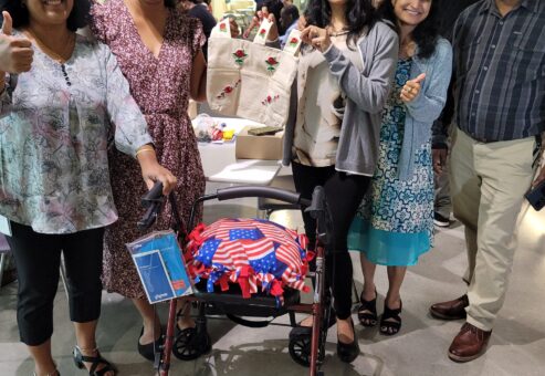 A group of five individuals, smiling and showing a thumbs up, stands around a rollator walker they have built. The walker features an American flag-themed cushion attached to it. One of the women is holding up a canvas tote bag decorated with colorful designs. Various supplies and items are visible on the table behind them, contributing to the Rollator Walker Build team building event.