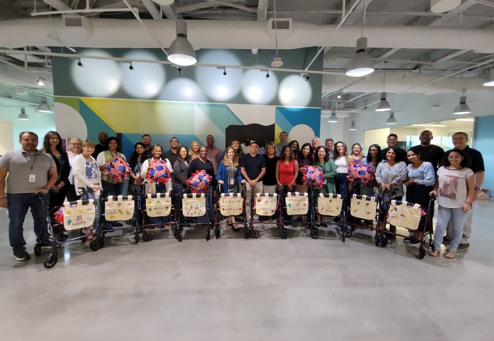 A group of participants stand behind a row of assembled rollator walkers, each adorned with decorated canvas bags and colorful fleece blankets, showcasing their completed project accomplished during a charitable team building program.