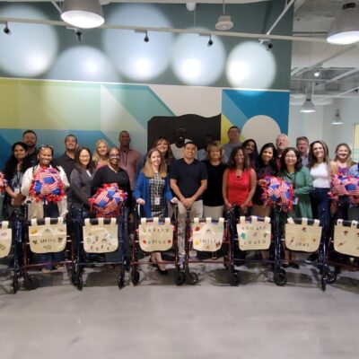 A group of participants stand behind a row of assembled rollator walkers, each adorned with decorated canvas bags and colorful fleece blankets, showcasing their completed project accomplished during a charitable team building program. thumbnail