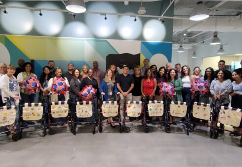A group of participants stand behind a row of assembled rollator walkers, each adorned with decorated canvas bags and colorful fleece blankets, showcasing their completed project accomplished during a charitable team building program.