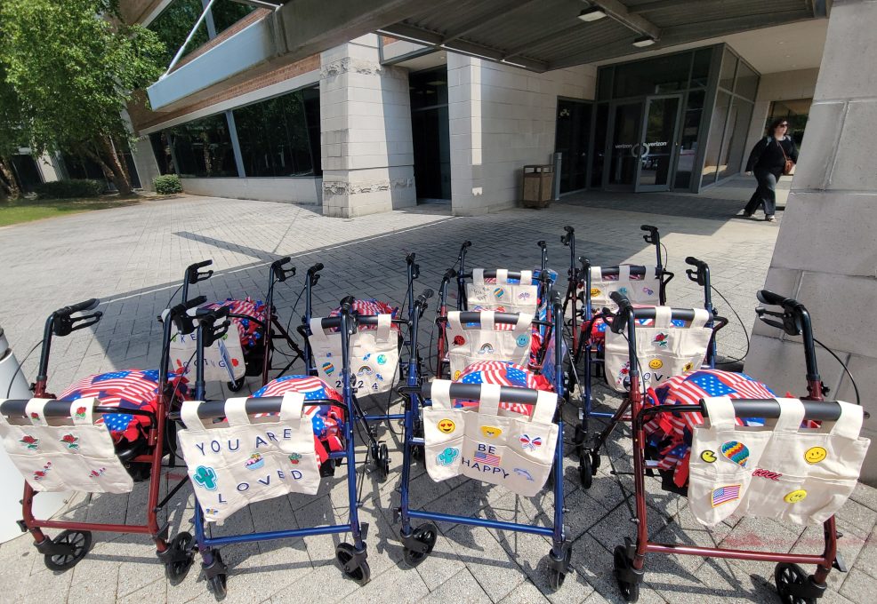 Group of assembled rollator walkers, decorated with personalized tote bags and American flag-themed pillows, lined up outdoors to showcase the result of a corporate charitable team building event.