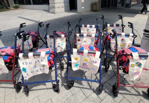Group of assembled rollator walkers, decorated with personalized tote bags and American flag-themed pillows, lined up outdoors to showcase the result of a corporate charitable team building event.