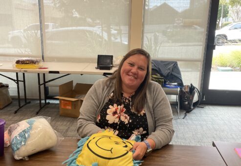 Smiling woman sits at a table holding a brightly colored no-sew pillow with a smiley face design, part of the Rollator Walker Build team building activity.