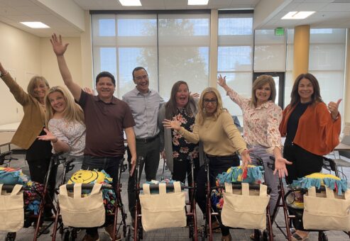 diverse group of eight adults joyfully posing behind their assembled rollator walkers, each adorned with a colorful fleece and a personalized accessory bag, showcasing their collaborative effort in a well-lit room.