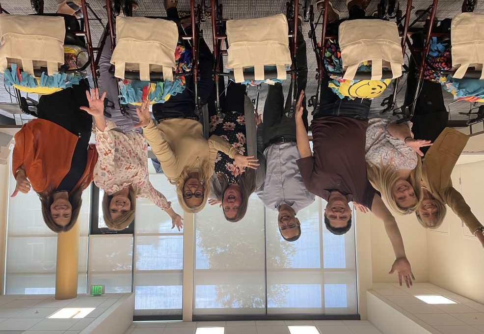 diverse group of eight adults joyfully posing behind their assembled rollator walkers, each adorned with a colorful fleece and a personalized accessory bag, showcasing their collaborative effort in a well-lit room.