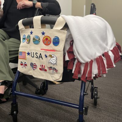 A rollator walker is decorated with a canvas bag displaying various USA-themed patches and a white and red fleece blanket draped over the seat. thumbnail