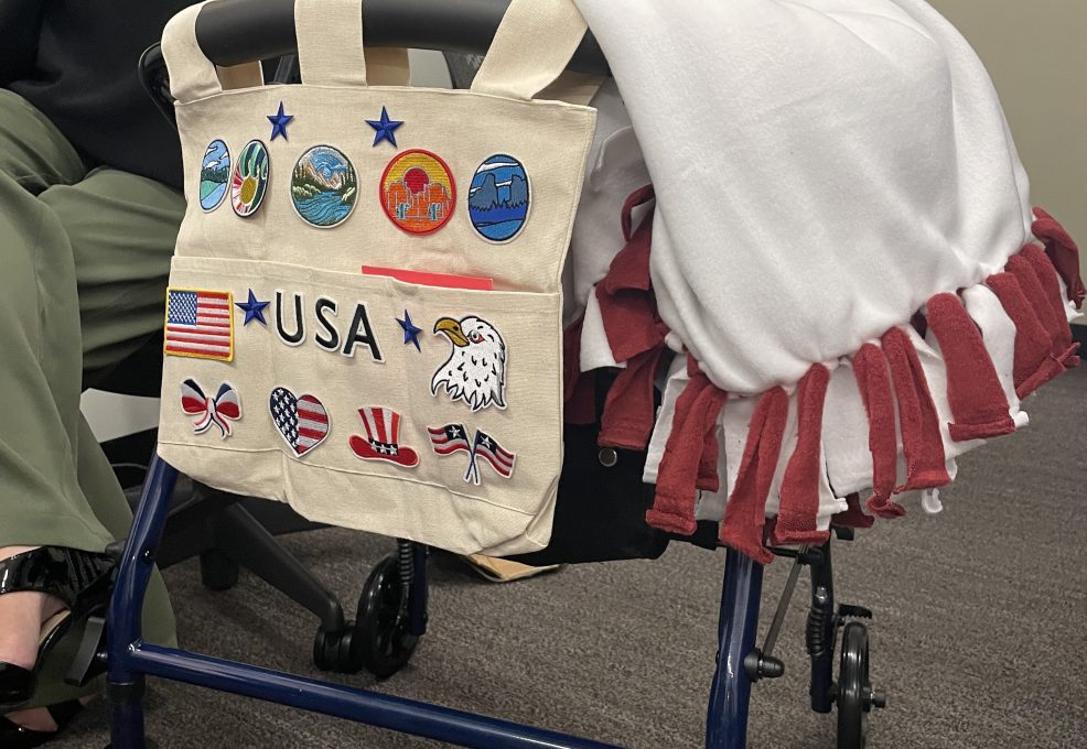 A rollator walker is decorated with a canvas bag displaying various USA-themed patches and a white and red fleece blanket draped over the seat.