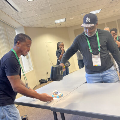 Two participants testing the performance of a small model car on a table during the STEM Speedway team building activity, with one participant using a fan to simulate wind conditions, showcasing teamwork, experimentation, and problem-solving. thumbnail