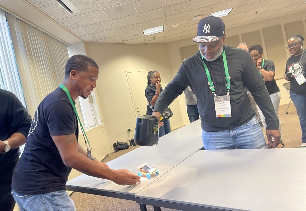 Two participants testing the performance of a small model car on a table during the STEM Speedway team building activity, with one participant using a fan to simulate wind conditions, showcasing teamwork, experimentation, and problem-solving.