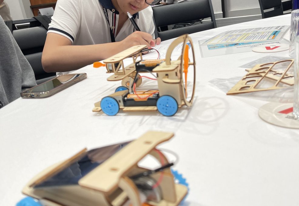 A participant focused on assembling a miniature car from a STEM kit during the STEM Speedway team building activity, with completed wooden model cars on the table, highlighting precision, creativity, and hands-on learning.