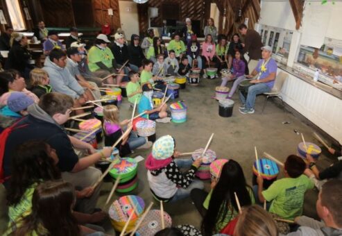 Josh Robinson leads a large group of children and adults in an interactive drumming circle. Participants, seated in a large wooden room, play colorful drums as Josh guides them through the activity, fostering a sense of rhythm, teamwork, and community engagement.