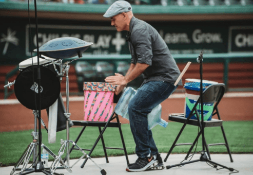 Josh Robinson performs a dynamic drumming session at an outdoor venue, playing various percussion instruments. Wearing a casual outfit and cap, he engages with the drums set up on chairs, delivering an energetic and interactive performance for the audience.