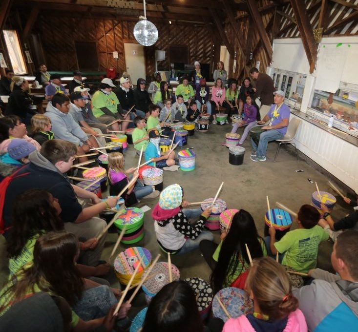 Josh Robinson leads a large group of children and adults in an interactive drumming circle. Participants, seated in a large wooden room, play colorful drums as Josh guides them through the activity, fostering a sense of rhythm, teamwork, and community engagement.