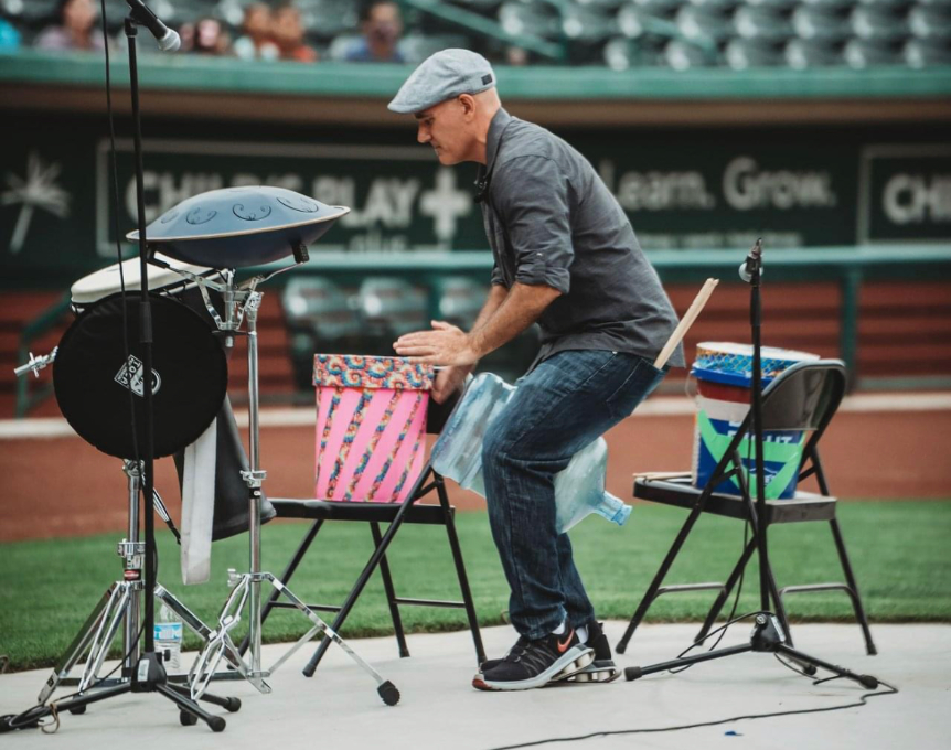 Josh Robinson performs a dynamic drumming session at an outdoor venue, playing various percussion instruments. Wearing a casual outfit and cap, he engages with the drums set up on chairs, delivering an energetic and interactive performance for the audience.