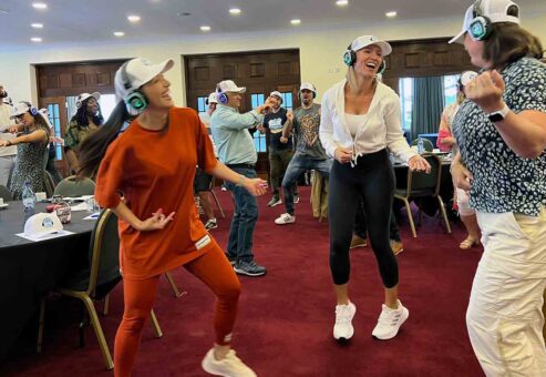 Participants in a Sound Crowd team building event laugh and dance while wearing noise-cancelling headphones. The group enthusiastically engages in the activity, which encourages non-verbal interaction through sound and movement, fostering fun and team connection.