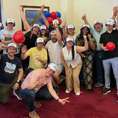 A cheerful group of Sound Crowd participants wearing white caps and headphones pose with balloons and enthusiastic gestures, celebrating the successful conclusion of their team building event. The atmosphere is energetic, and everyone looks engaged and happy. thumbnail