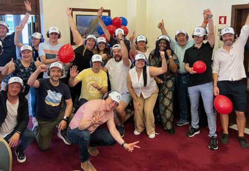 A cheerful group of Sound Crowd participants wearing white caps and headphones pose with balloons and enthusiastic gestures, celebrating the successful conclusion of their team building event. The atmosphere is energetic, and everyone looks engaged and happy.