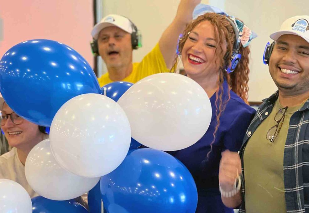 A lively group of Sound Crowd participants smiling and holding blue and white balloons while wearing white caps and headphones during a team building event.