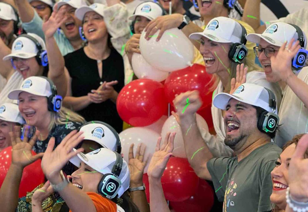 A large group of Sound Crowd participants cheering and smiling while wearing white caps and headphones. They are enthusiastically raising their hands, and red and white balloons are visible in the background, adding to the festive and vibrant atmosphere of the team building event.