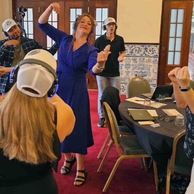 Participants at the Sound Crowd team building event dance and groove while wearing white caps and blue headphones. One woman in a royal blue dress is leading the group with expressive dance movements, while others follow along, all enjoying the music on their headsets. thumbnail
