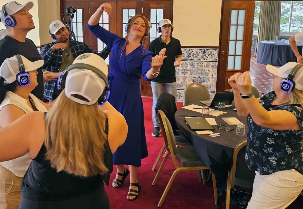 Participants at the Sound Crowd team building event dance and groove while wearing white caps and blue headphones. One woman in a royal blue dress is leading the group with expressive dance movements, while others follow along, all enjoying the music on their headsets.