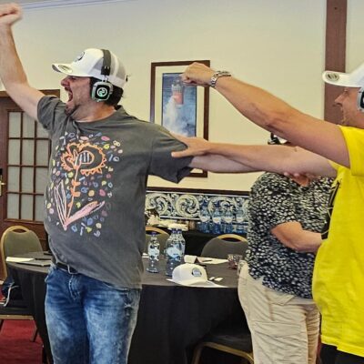Two participants in a Sound Crowd team building event enthusiastically dance while wearing white caps and headphones. One man in a graphic t-shirt and jeans throws his arms up in excitement, while the other, wearing a yellow t-shirt, joins in with equally energetic movements. thumbnail