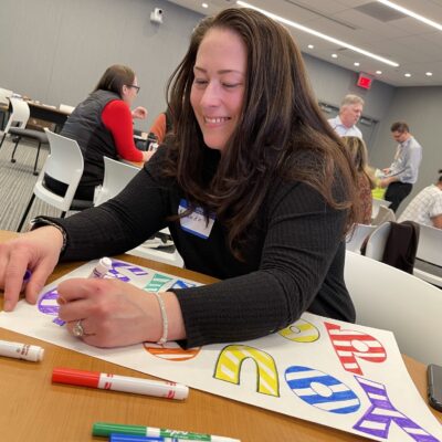 A participant joyfully colors a holiday-themed sign as part of the Hope for the Holidays team building event, creating personalized gift baskets for seniors in local nursing homes. thumbnail