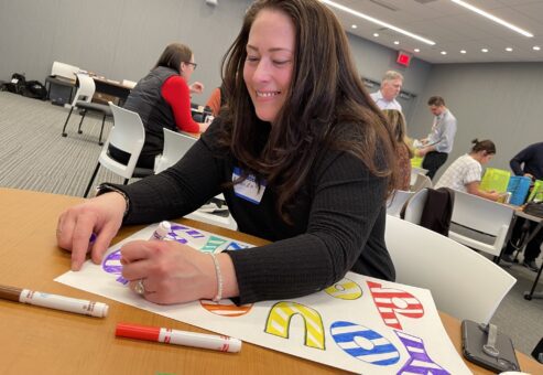 A participant joyfully colors a holiday-themed sign as part of the Hope for the Holidays team building event, creating personalized gift baskets for seniors in local nursing homes.