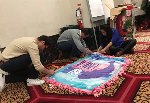 Team members collaborate during the Hope for the Holidays event, carefully crafting a fleece blanket to donate to local seniors as part of their charitable efforts.