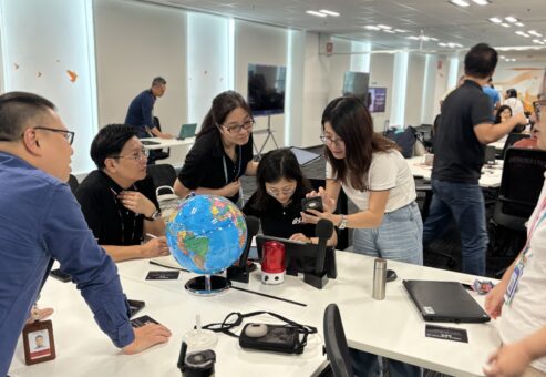 A team collaborates in a team building AI-powered news broadcast challenge, brainstorming and working together on roles like scriptwriting, research, and production.