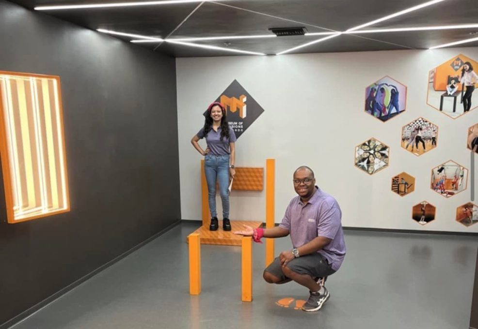 Team Pink posing in the Museum of Illusions' 'Nobody Puts Baby in the Corner' exhibit during a scavenger hunt. The optical illusion creates a fun and interactive challenge for the team to explore and solve.
