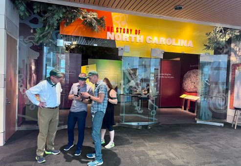 Team participating in a museum scavenger hunt at the North Carolina history exhibit. Using a mobile device, they collaborate to solve clues, enhancing teamwork, problem-solving, and engagement with culture.