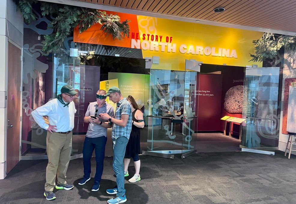 Team participating in a museum scavenger hunt at the North Carolina history exhibit. Using a mobile device, they collaborate to solve clues, enhancing teamwork, problem-solving, and engagement with culture.